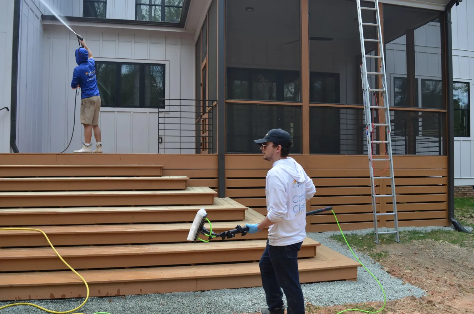 Two Carolina Sky technicians at work, one washing siding from the deck while the other carries a water-fed window brush