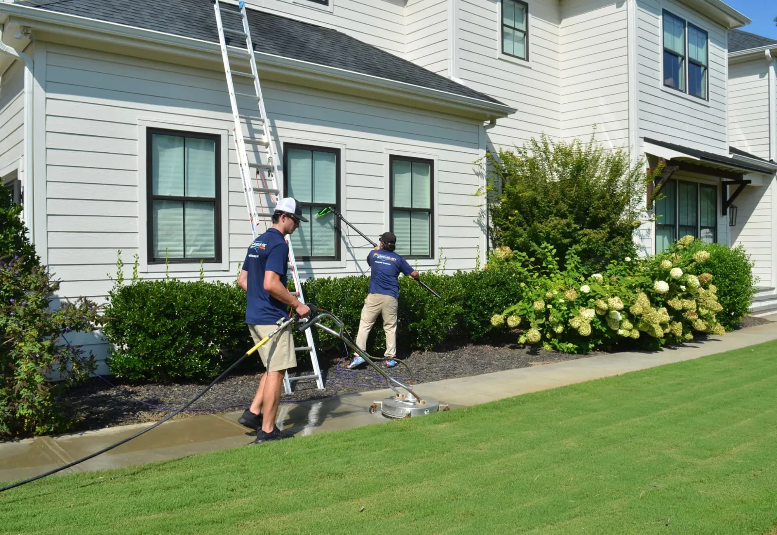 Two technicians cleaning a sidewalk and windows around a white home