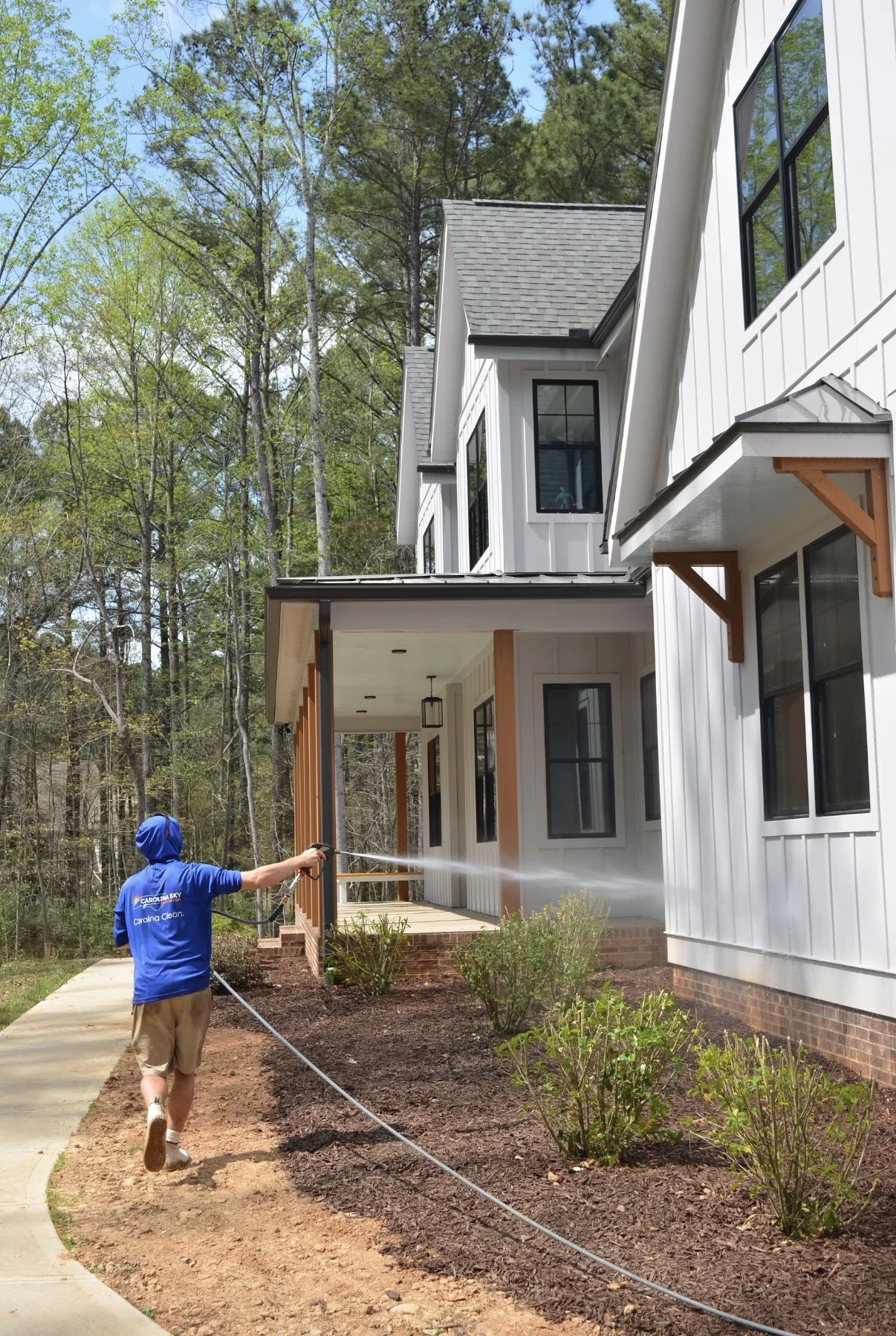 Technician soft washing the tall side wall of a white farmhouse